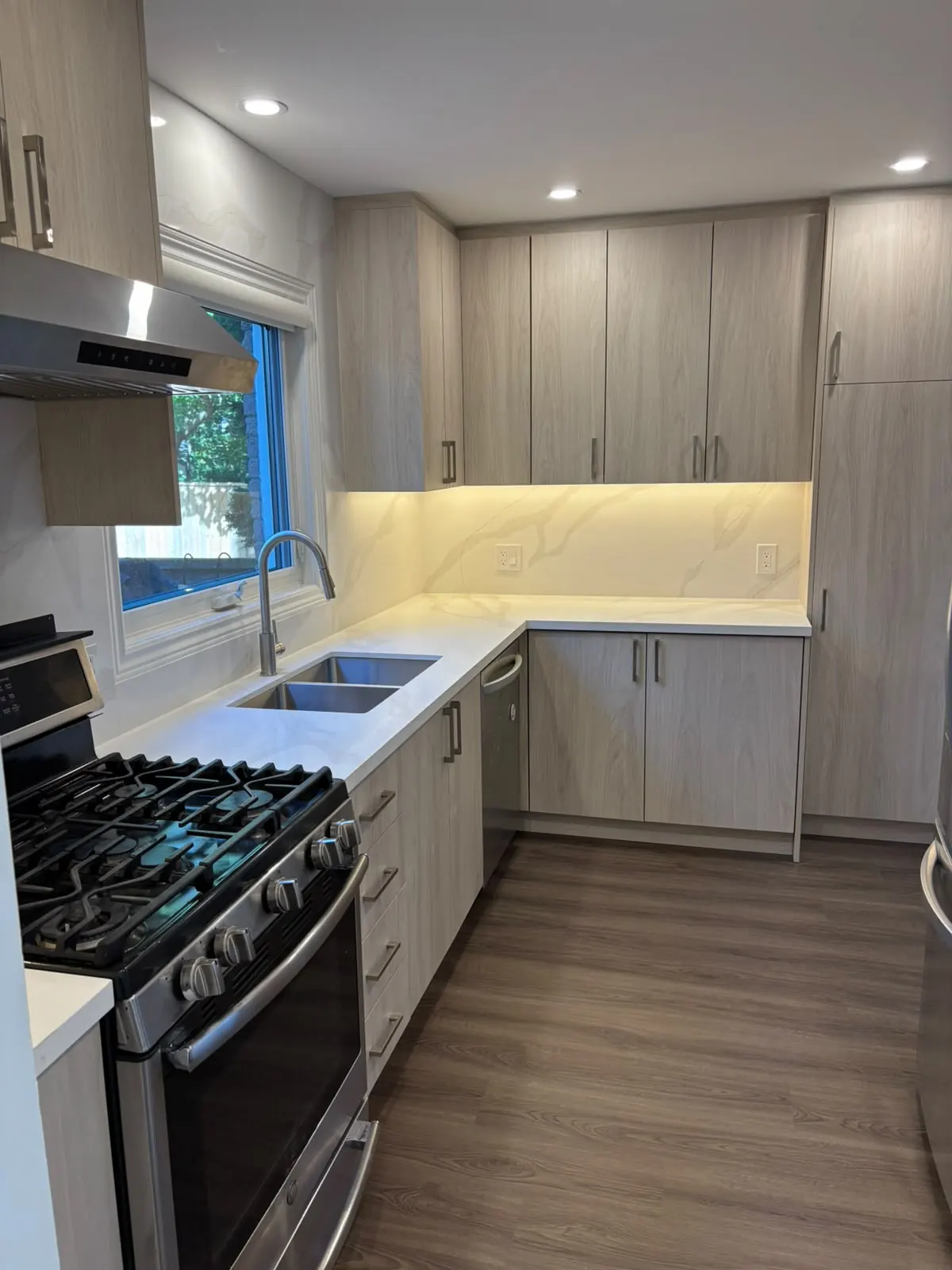 Renovated galley kitchen with light wood-grain cabinets, a white stone-look backsplash and a stainless gas range